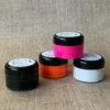 Vier kleine Farbdosen mit den Farben Schwarz, Neon-Orange, Neon-Pink und Weiß gefüllt auf beigem Hintergrund.