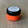 Kleine, runde und durchsichtige Dose mit schwarzem Declek gefüllt mit neon-orangener Textildruckfarbe. Ein weißer Inhalts-Aufkleber klebt auf dem Deckel.