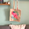 Eine Stoffbeutel mit langen Henkeln aus ungefärbter Baumwolle hängt an einem Regal. Die Vorderseite der Tasche ist abstrakt mit bunten Neonfarben bemalt, darüber sind schwarze Linien in einem abstrakten Muster gezeichnet. An einem Henkel ist ein türkisfarbiger Pompon-Anhänger mit Holzperle angebracht.