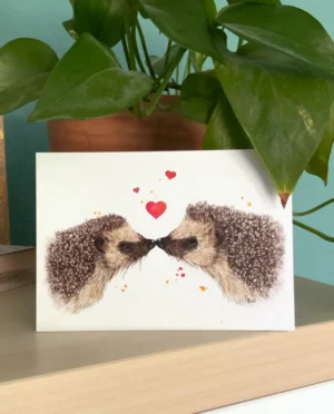 Eie Postkarte steht auf einem Regal vor einer Pflanze mit grünen Blättern. Aus der Karte sind zwei gezeichnete Igel, die sich an der Schnauze berühren, Herzchen steigen auf.