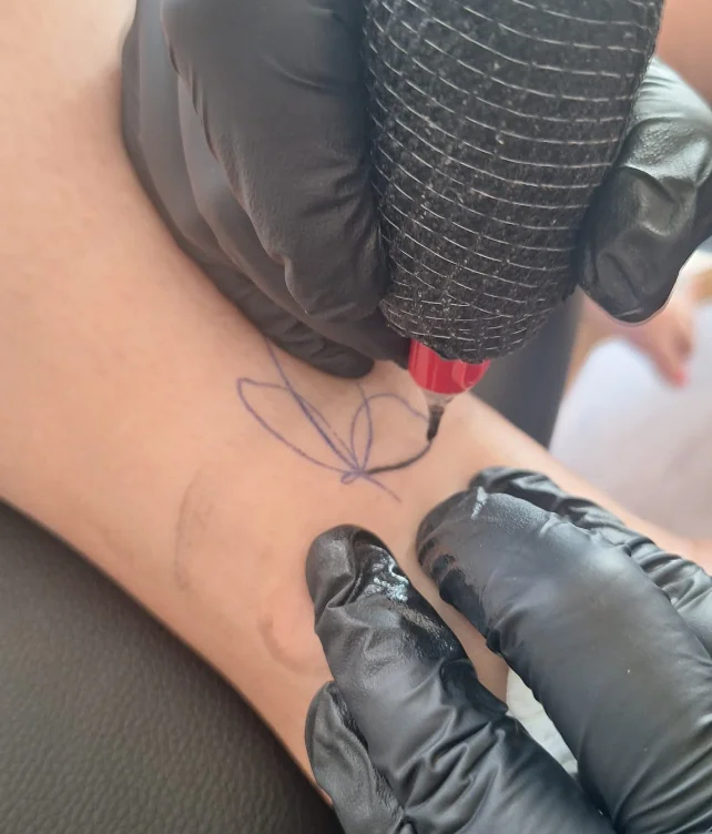 Beim Tätowieren - Hände in schwarzen Handschuhen halten eine Tattoomaschine und tätowieren einen Schmetterling auf ein Bein.